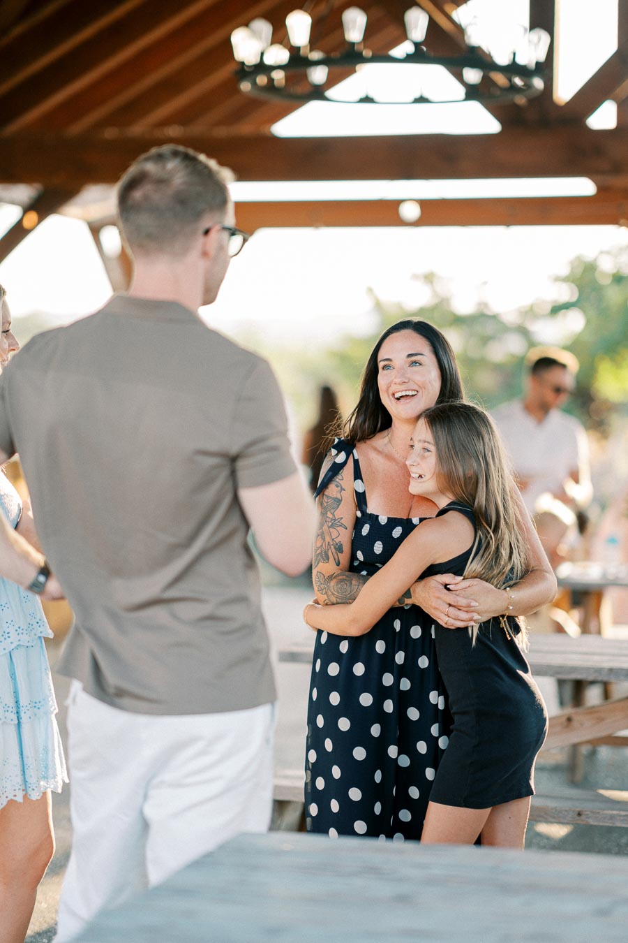 Mother and daughter hugging at an outdoor gathering, under a wooden pavilion, with people socializing in the background.