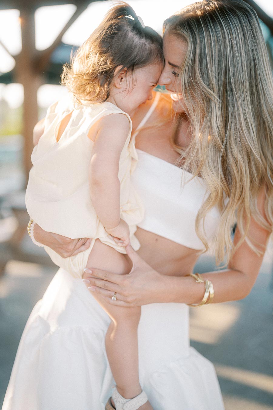 A smiling woman holding a baby close in a warm embrace, both in light-colored outfits, enjoying a sunny day outdoors.