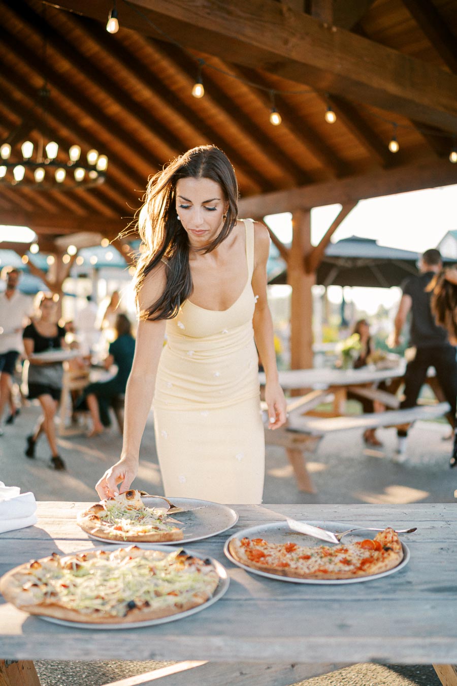 Woman in a yellow dress selecting pizza from a wooden table at an outdoor gathering, with people and string lights in the background.