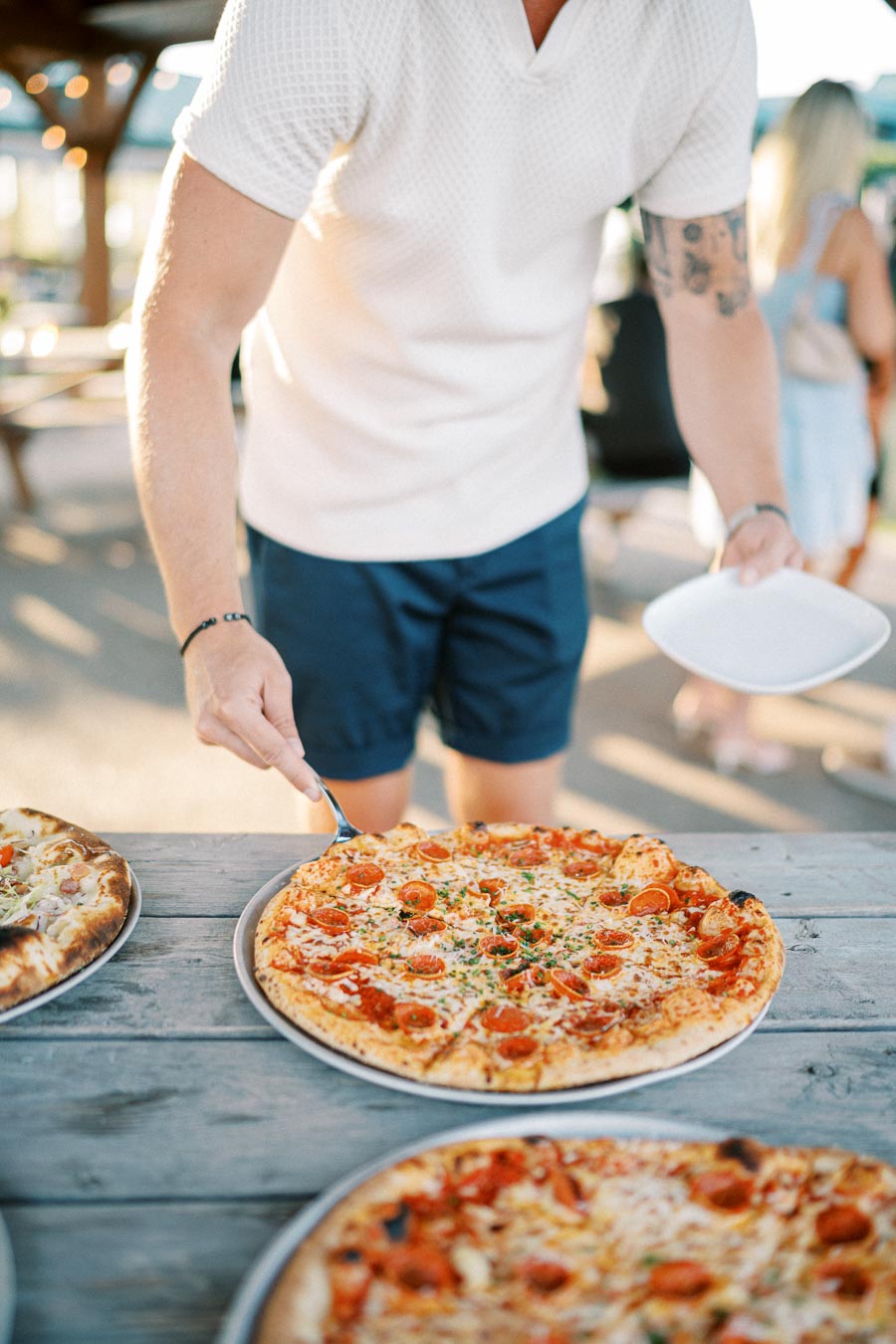A person in a white shirt and blue shorts serves a slice of pepperoni pizza outdoors on a wooden table, with other pizzas visible nearby.