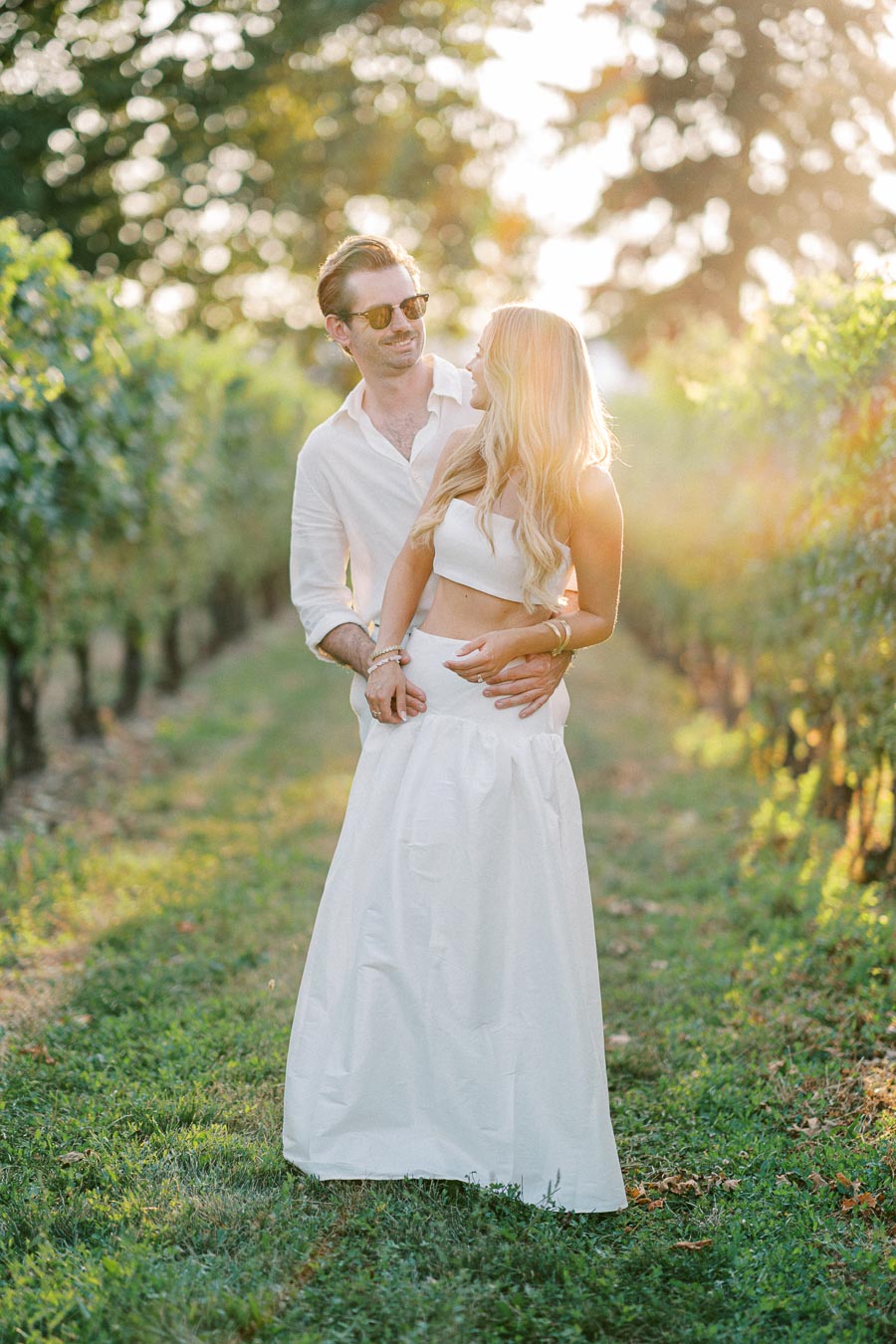 A couple embracing in a sunlit vineyard, wearing stylish white outfits, with greenery in the background, capturing a romantic and serene moment.
