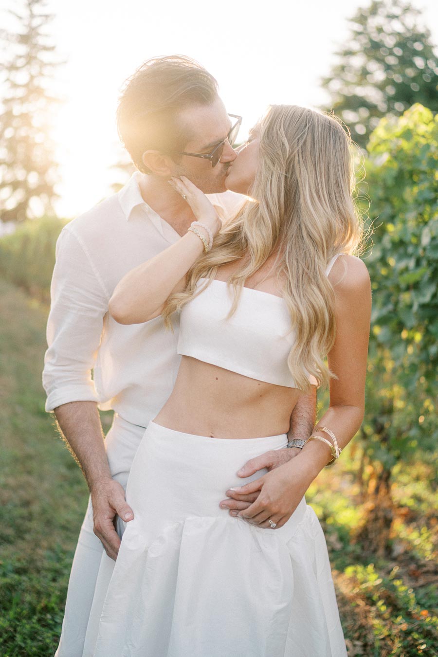 A couple kissing in a sunlit vineyard, both dressed in white, with lush greenery in the background.