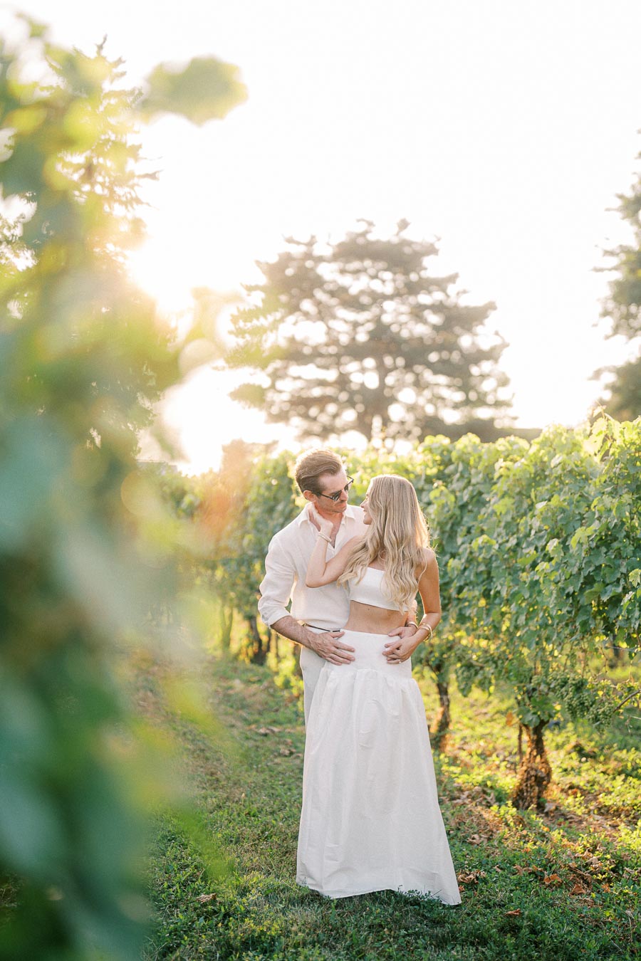 Romantic couple embracing amidst lush vineyard with sunlight filtering through trees, capturing a serene and intimate moment, ideal for wedding inspiration or engagement photography in a natural setting.
