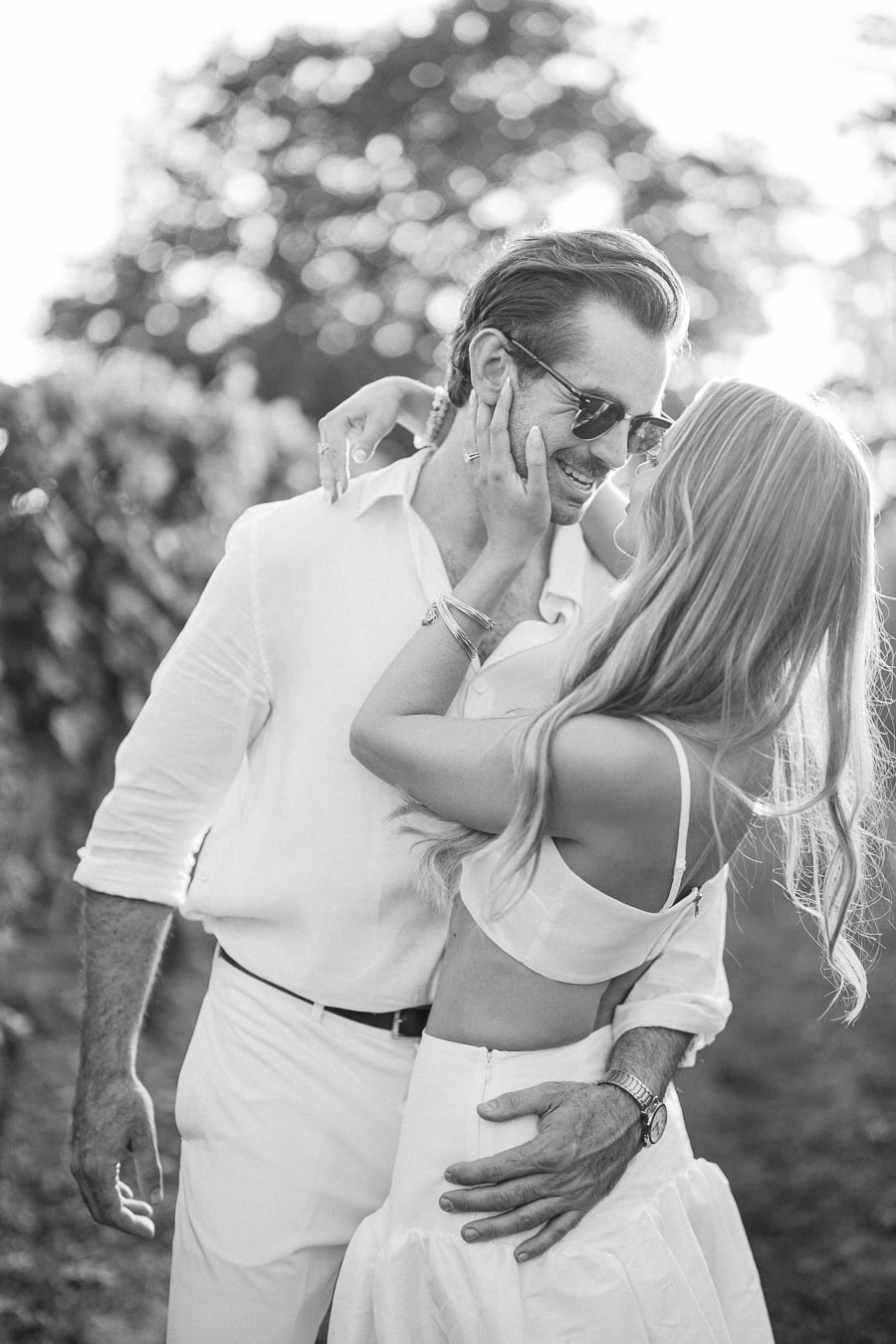 A couple embracing in a sunlit vineyard, wearing casual white outfits, capturing a romantic moment in black and white photography.