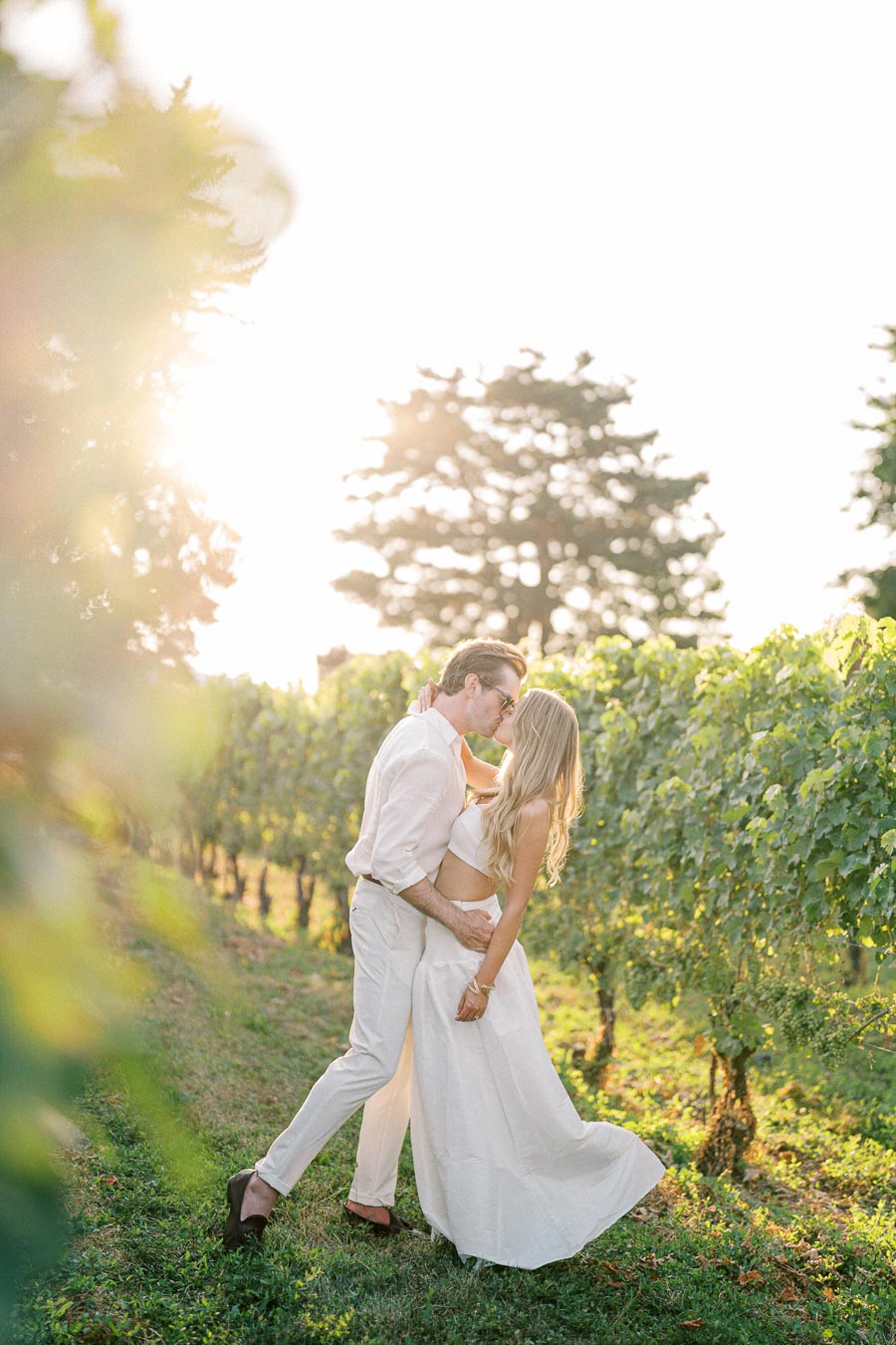 A couple dressed in white kisses passionately in a sunlit vineyard, surrounded by lush green vines, during golden hour.