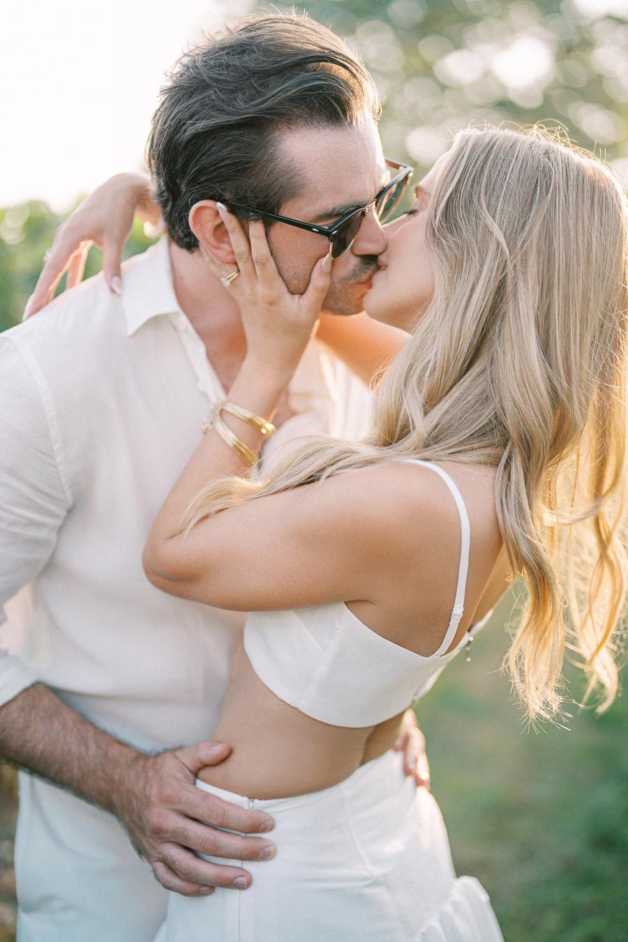 A couple sharing a romantic kiss in a sunny outdoor setting, both dressed in white outfits, surrounded by a soft, warm glow and blurred greenery in the background.