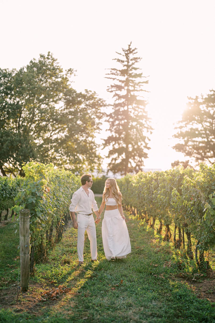 A couple in white clothing walking hand in hand through a lush vineyard at sunset, surrounded by green grapevines and warm, golden sunlight.