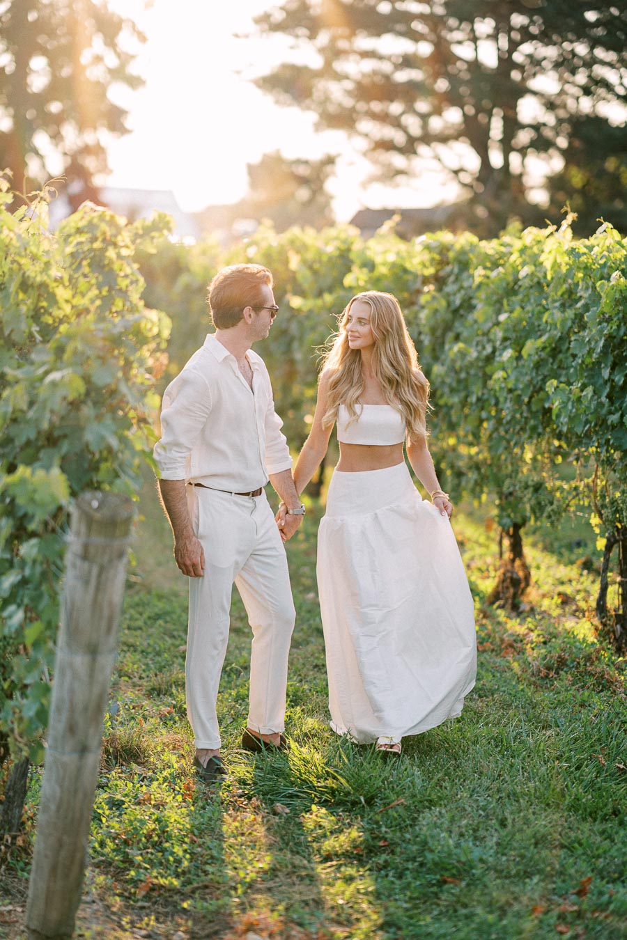 A romantic couple walking through a sunlit vineyard, dressed in stylish white outfits, surrounded by lush green grapevines and a soft, golden glow from the setting sun.