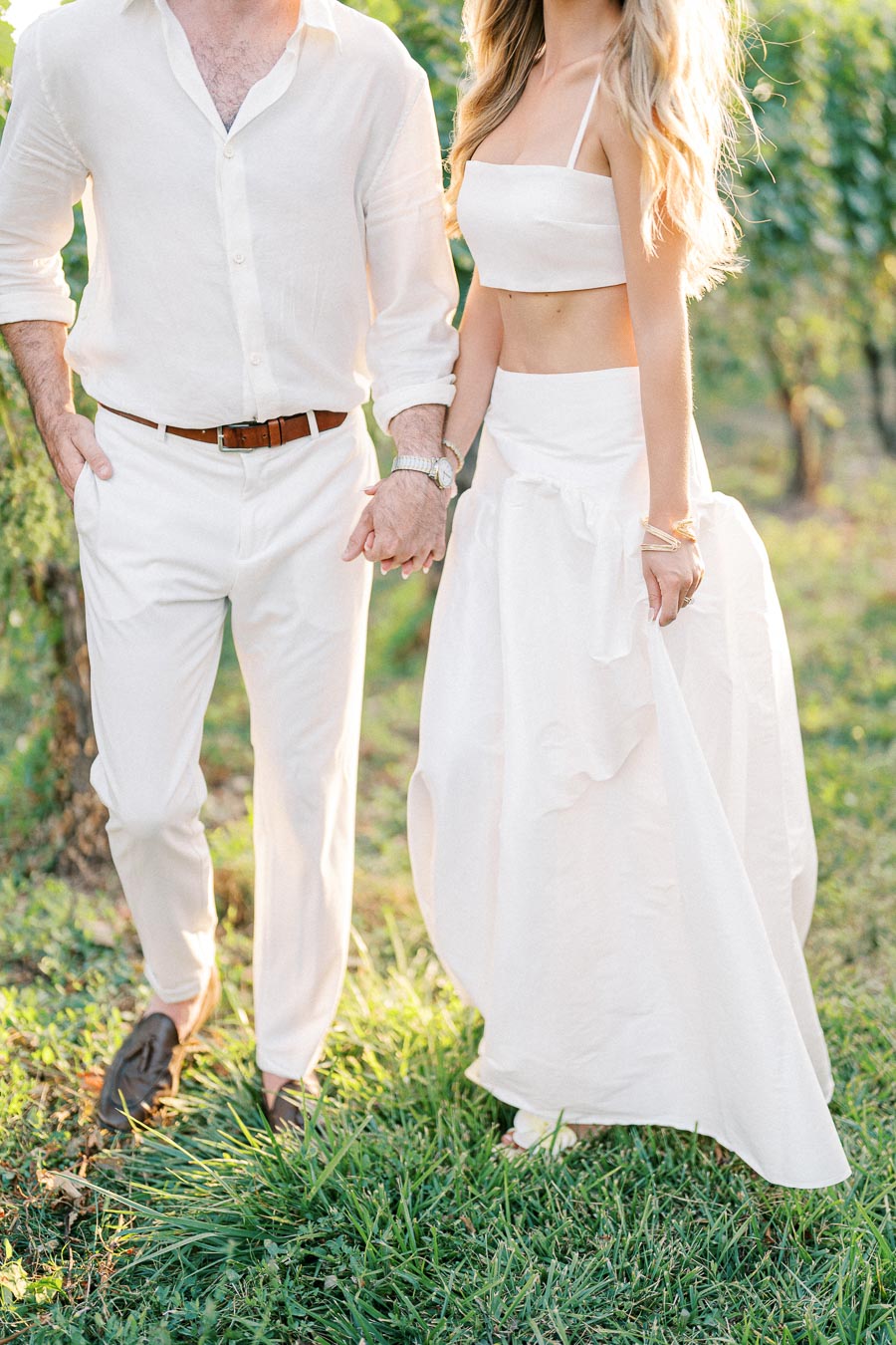 A couple dressed in elegant white outfits holding hands while walking through a sunlit vineyard, symbolizing romance and tranquility in nature.