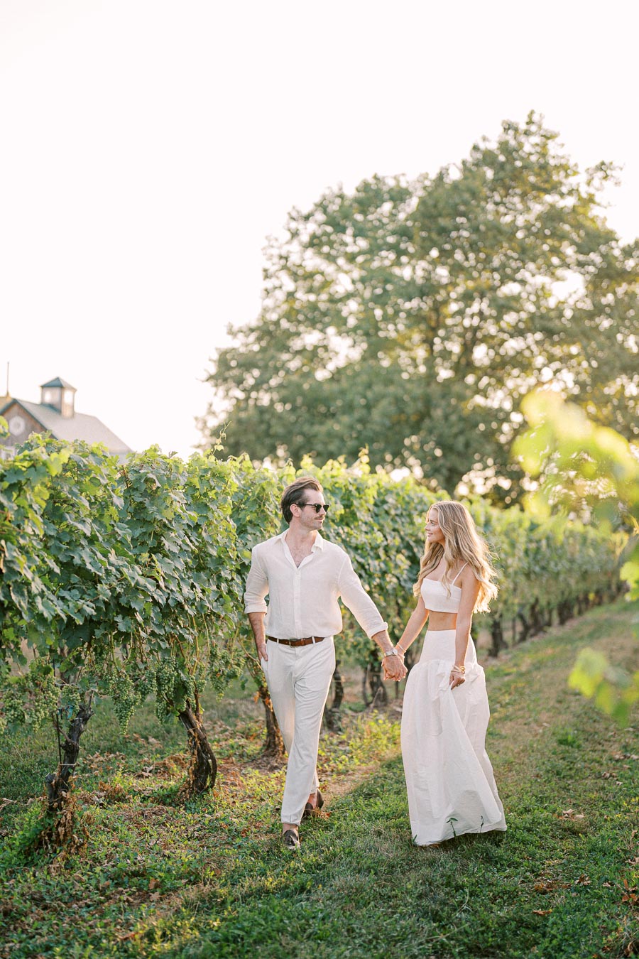 A couple walking hand in hand through a vineyard at sunset, wearing white outfits; lush green vines on both sides create a romantic and serene atmosphere perfect for a summer day.