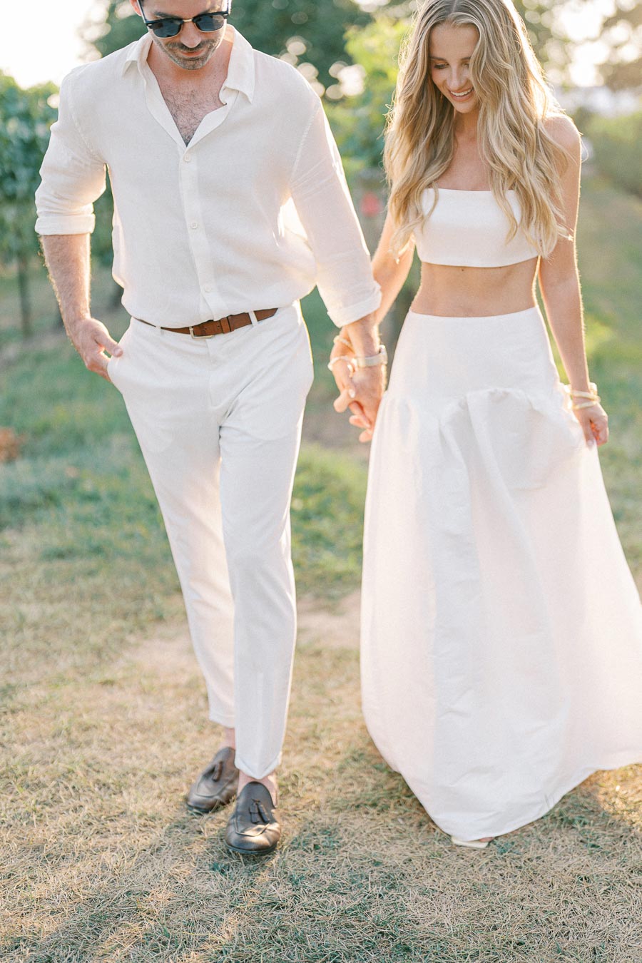 A couple dressed in stylish, coordinated white outfits walking hand in hand in a sunlit outdoor setting, surrounded by greenery, conveying a sense of romance and elegance.