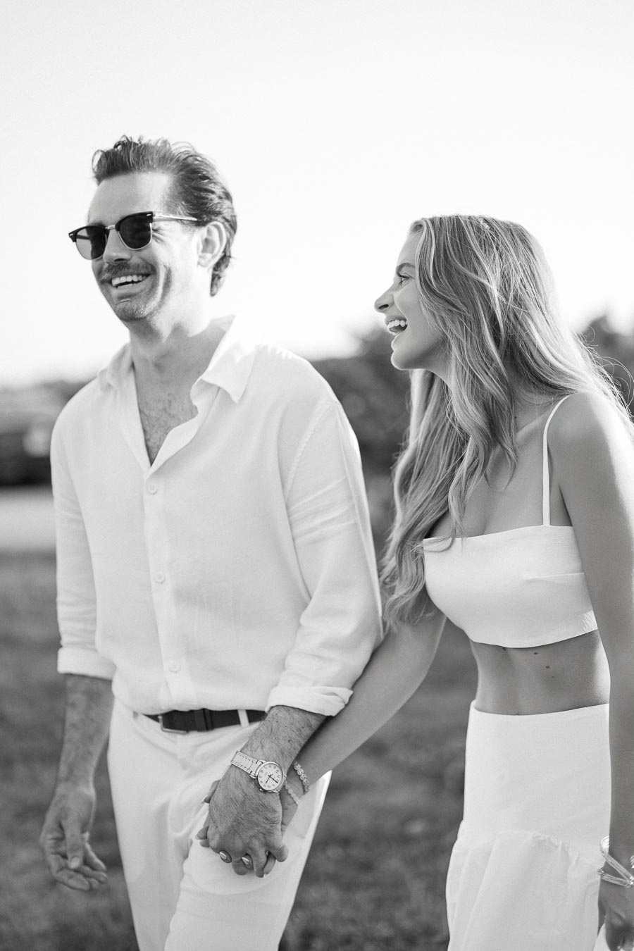A couple laughing and holding hands, walking outdoors in stylish white clothing, exuding happiness and love.