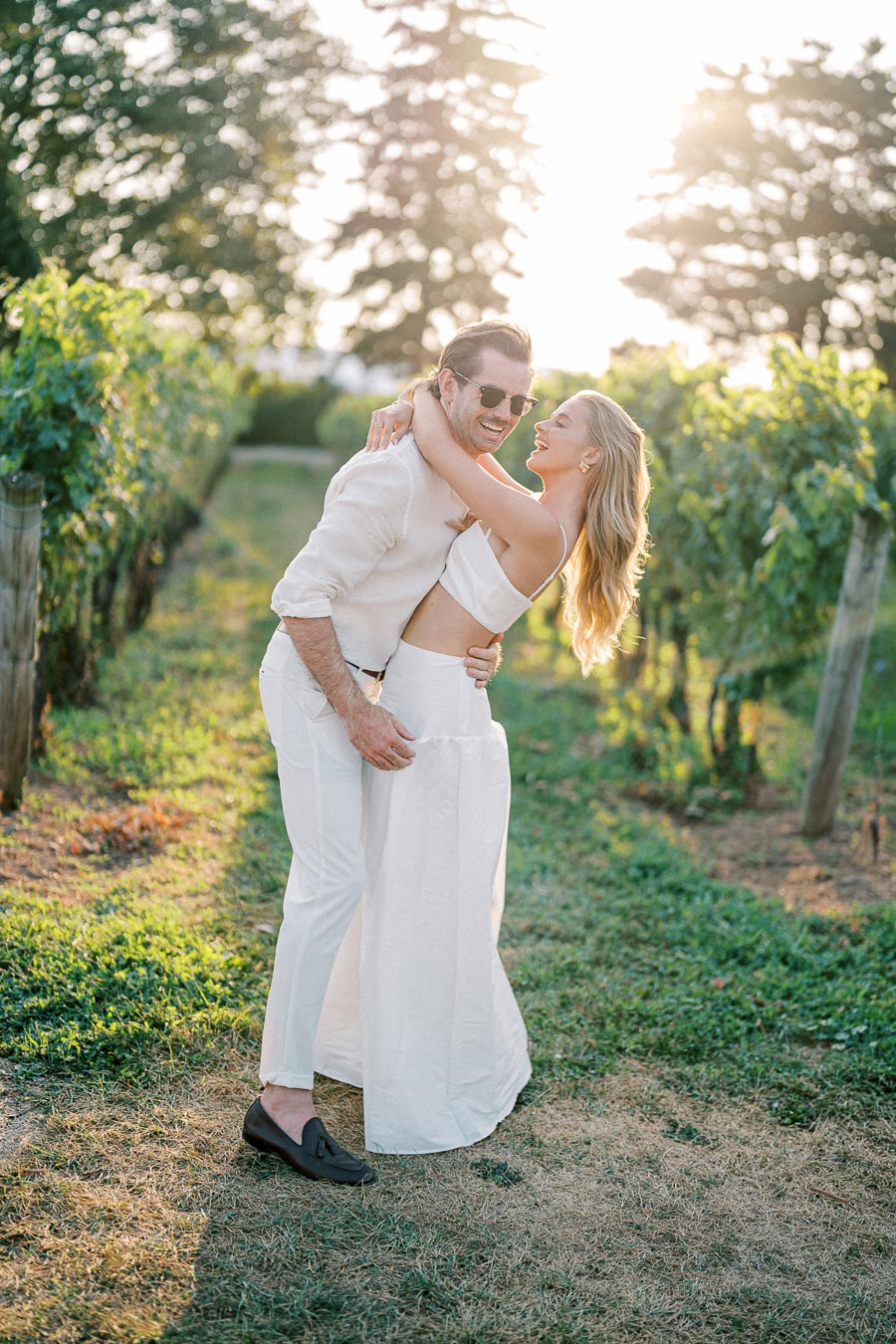 A couple in stylish white outfits embracing and laughing in a sunlit vineyard, surrounded by lush green grapevines.