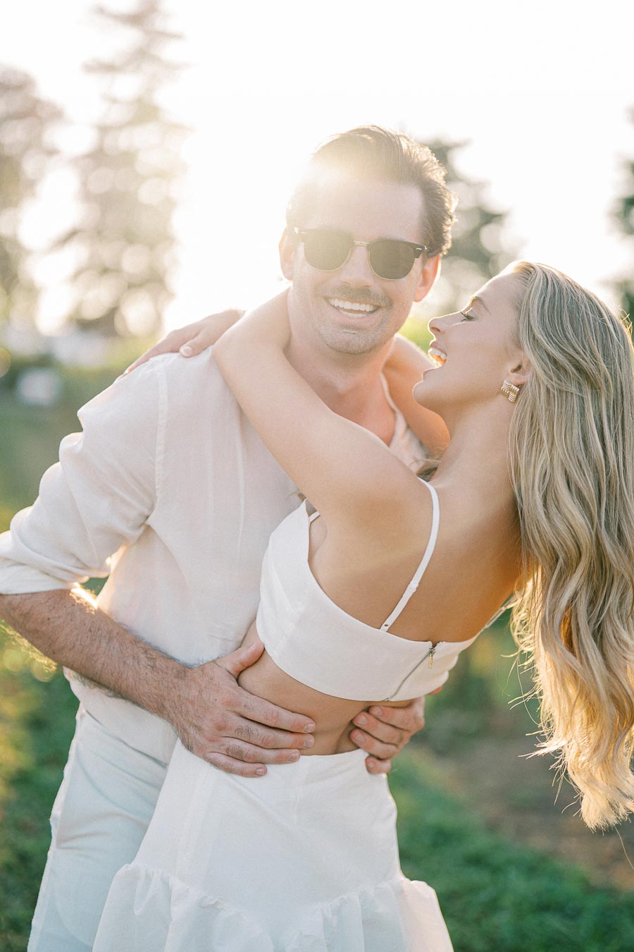 A couple embracing and smiling outdoors in a sunlit garden, both wearing white outfits, with soft focus trees in the background.
