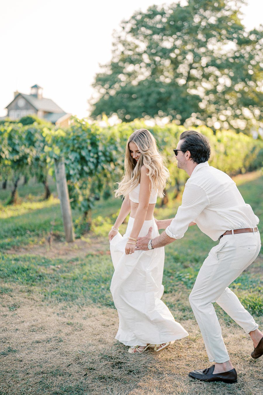 A couple playfully enjoying a sunny day in a vineyard, dressed in white summer attire, with lush green grapevines and a rustic building in the background.