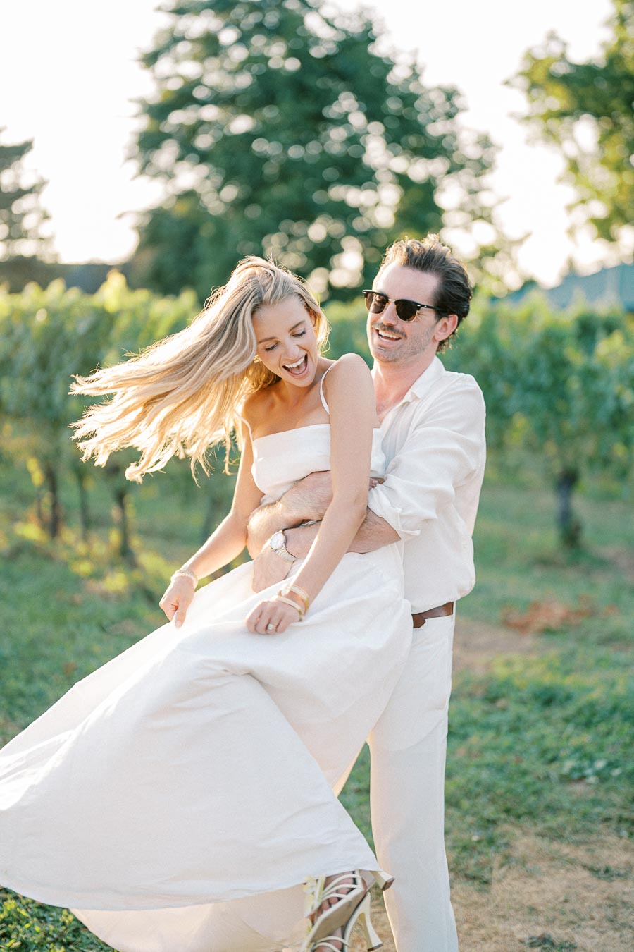 A joyful couple in white clothing enjoying a summer day in a vineyard, with the man playfully lifting the woman as she laughs, surrounded by lush green vines and soft sunlight.