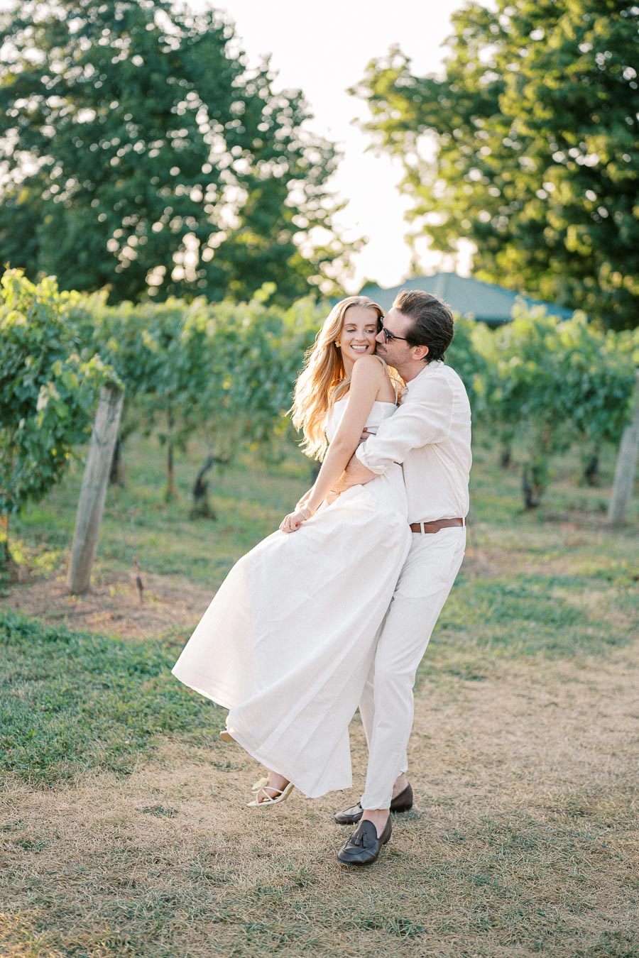 A couple in white outfits joyfully embraces in a lush vineyard, surrounded by vibrant greenery on a sunny day.