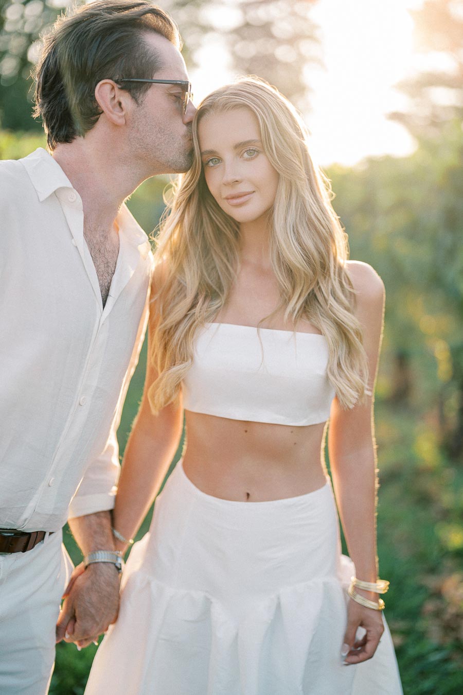 A couple dressed in white, standing in a sunlit vineyard; a man kisses a woman's forehead as they hold hands, exuding romance and style.