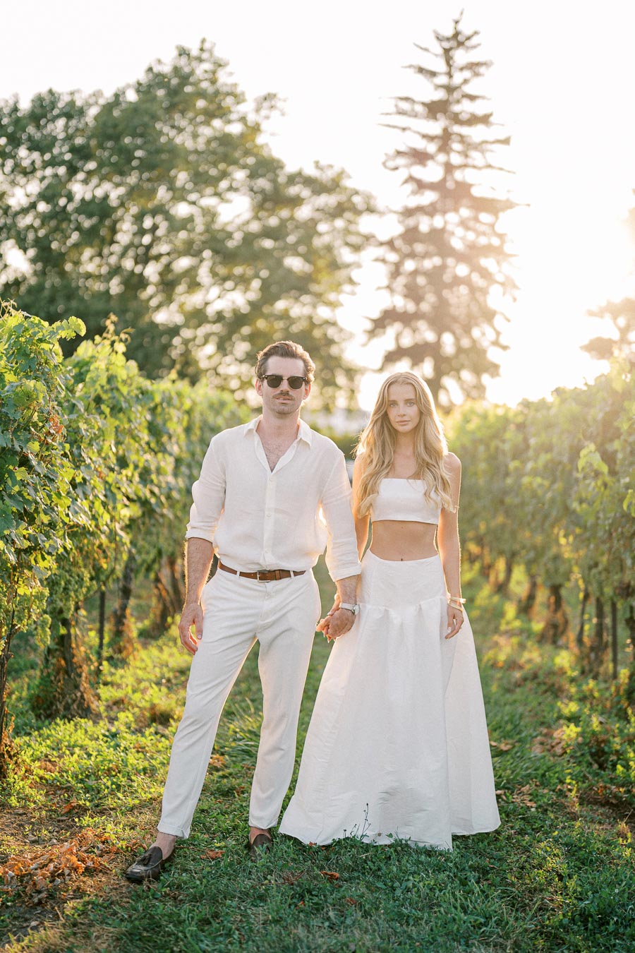 Couple in white clothing holding hands in a sunny vineyard.
