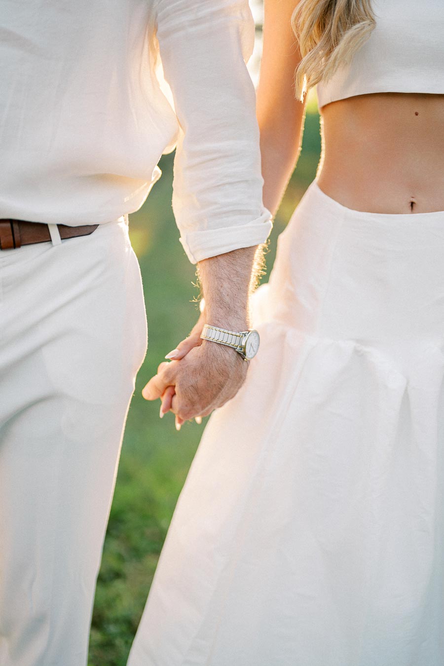 A couple holding hands at an outdoor event while dressed in light-colored clothing, highlighting a sense of togetherness and romance in a serene setting.