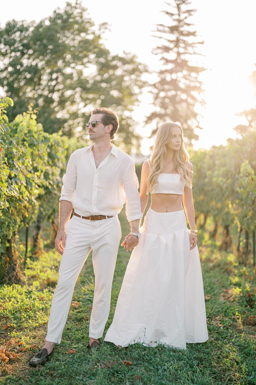 A couple in coordinated white outfits walking hand-in-hand through a sunlit vineyard, with lush green vines and trees in the background.