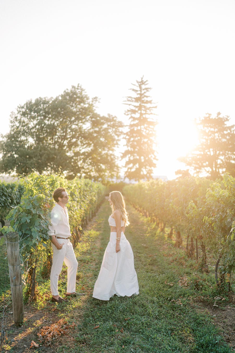 A couple in elegant white attire enjoying a sunny day in a lush vineyard, with rows of grapevines and trees in the background, capturing a romantic and serene moment.