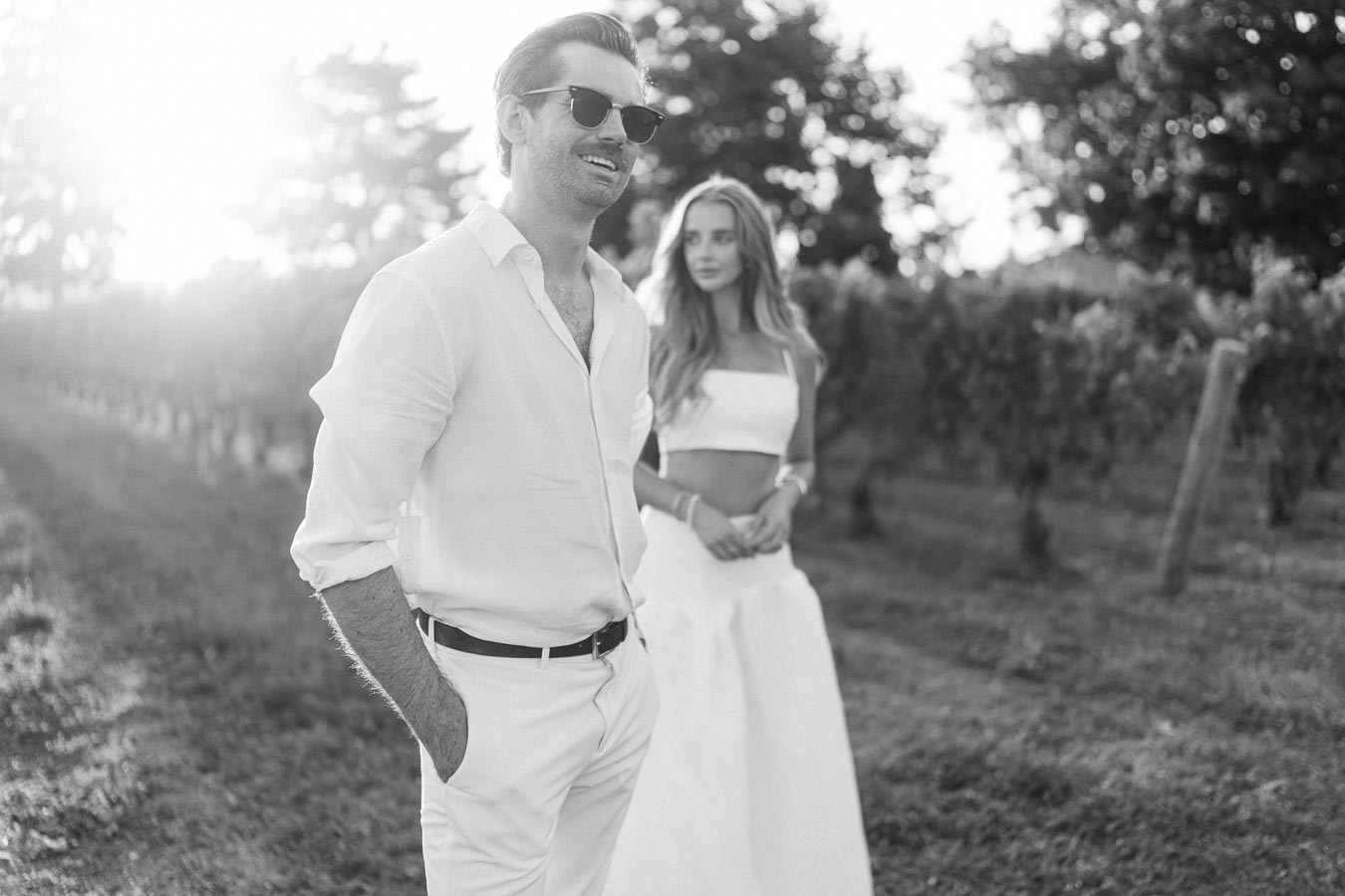 Stylish couple in elegant white outfits walking through a scenic vineyard in black-and-white photography, capturing a sunlit romantic moment.
