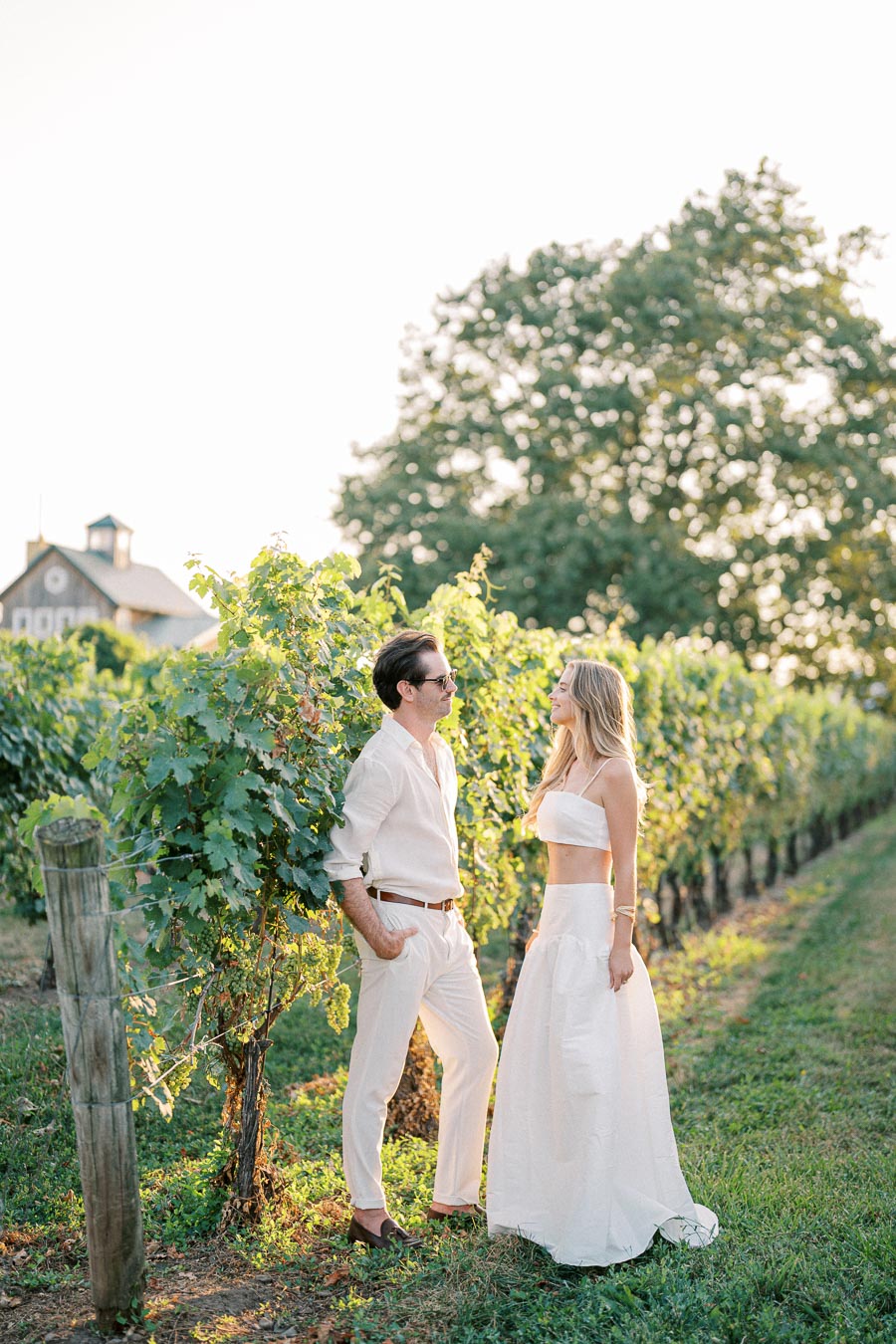 A couple in stylish white attire stands among lush green vineyard rows during sunset, with a rustic barn in the background, exuding a romantic and elegant atmosphere.