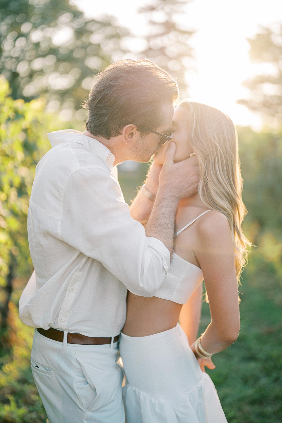 A couple kissing passionately in a sunlit vineyard, both dressed in white summer outfits, surrounded by lush greenery and warm evening light.