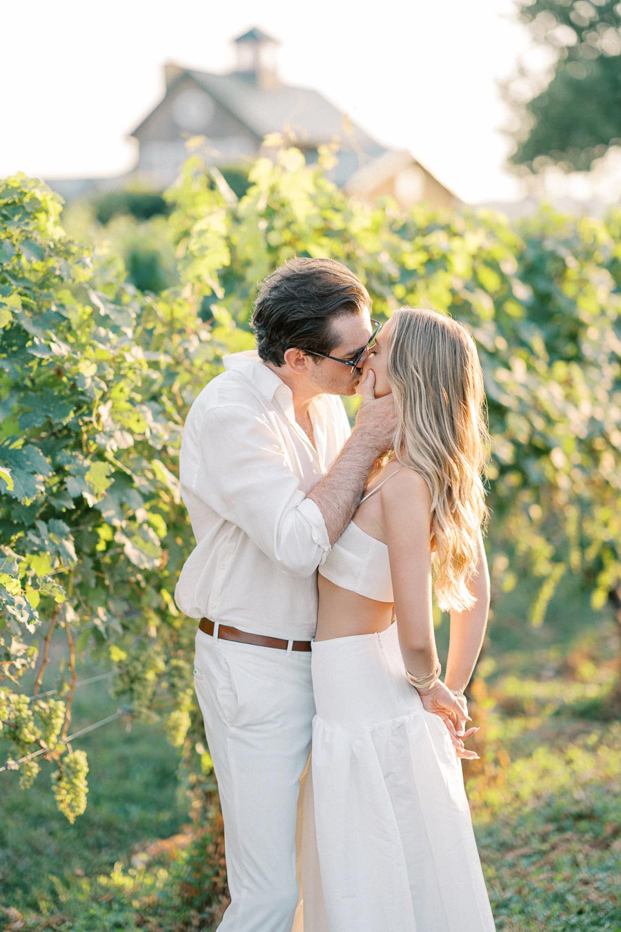 A romantic couple kissing in a sunlit vineyard, both dressed in white, with lush green grapevines and a blurred rustic building in the background.