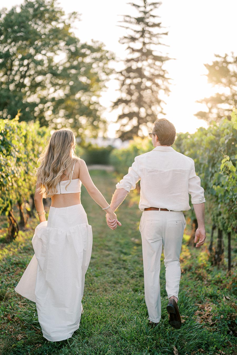 A couple holding hands walking through a vineyard at sunset, both wearing white outfits.