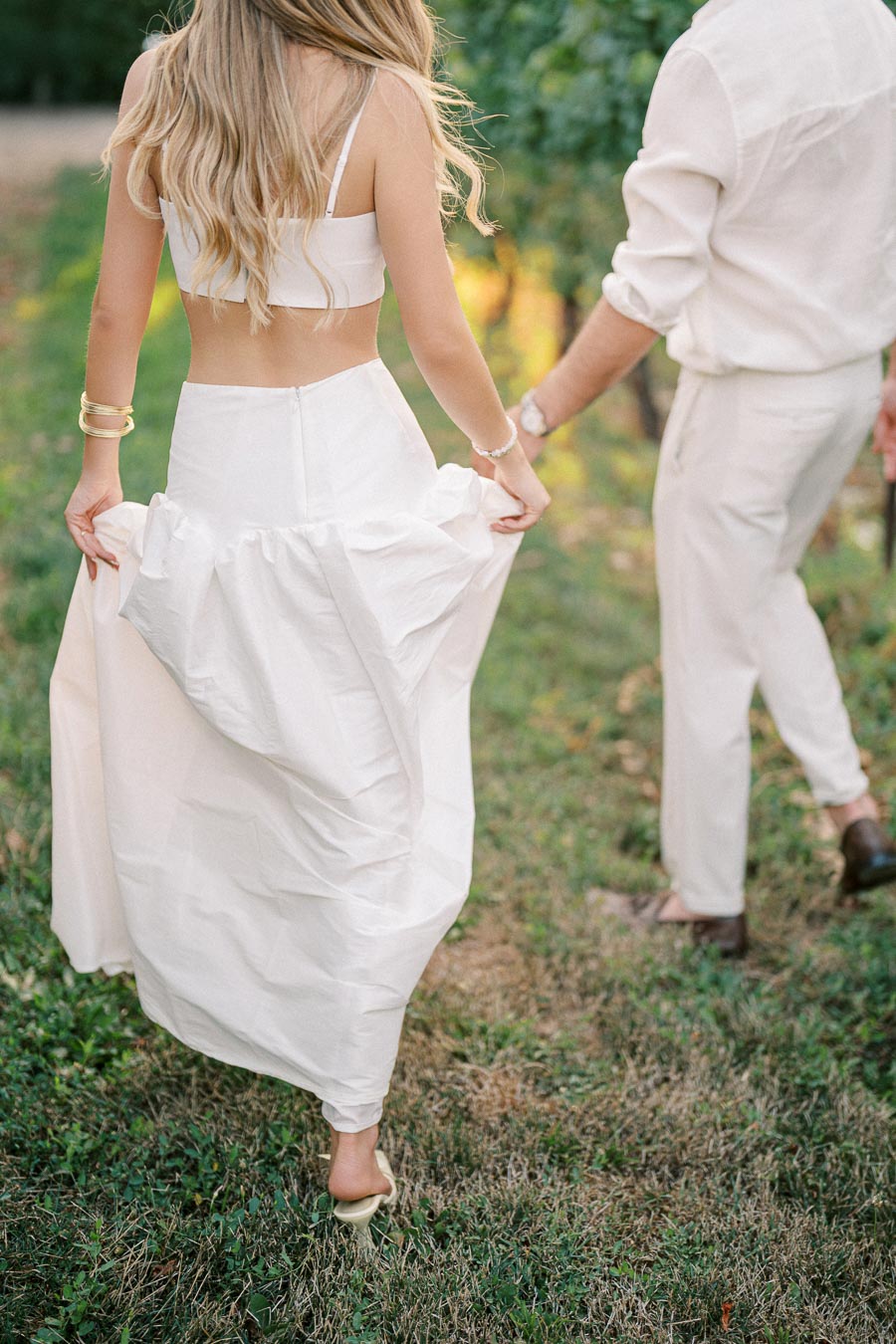 Couple walking hand in hand through a garden, woman in a white dress holds the hem, creating a romantic and serene atmosphere.