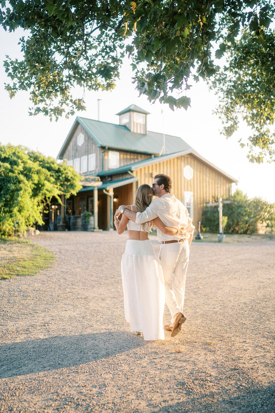 A couple walking arm in arm towards a rustic barn under a tree, wearing summer clothing and bathed in warm sunlight.