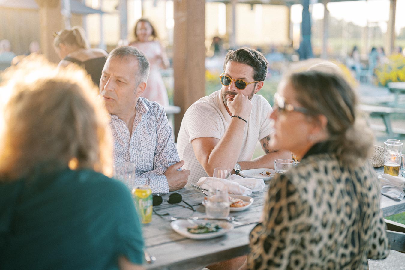 A group of people enjoy an outdoor dinner gathering at a rustic venue with warm sunlight illuminating the scene. Plates and drinks are on the wooden table, creating a relaxed and social atmosphere.
