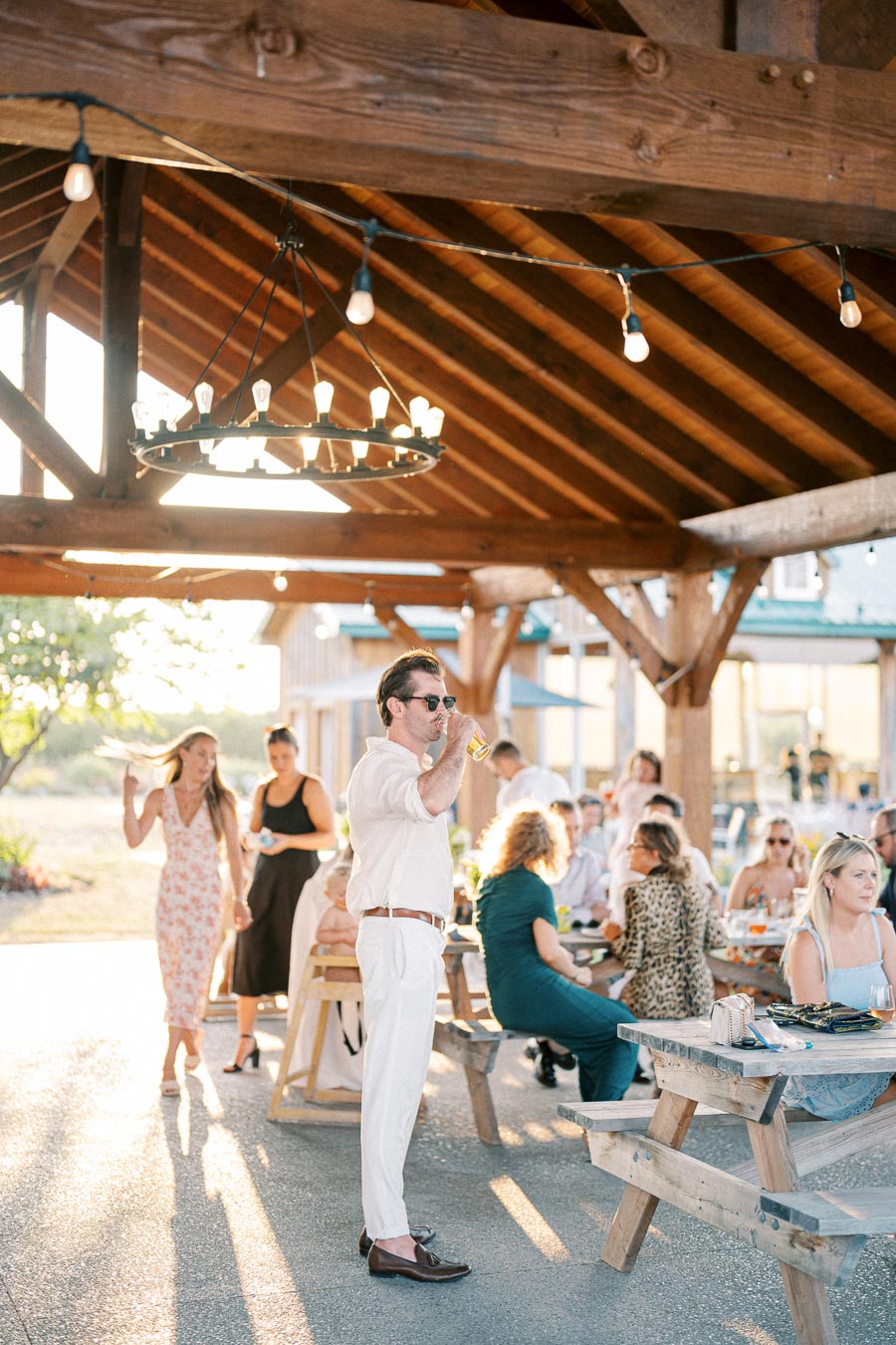 Outdoor gathering under a wooden pavilion with guests enjoying food and drinks; a man in white attire is standing and sipping a beverage while others are seated at wooden picnic tables. Warm lighting and rustic decor create a relaxed and festive atmosphere.