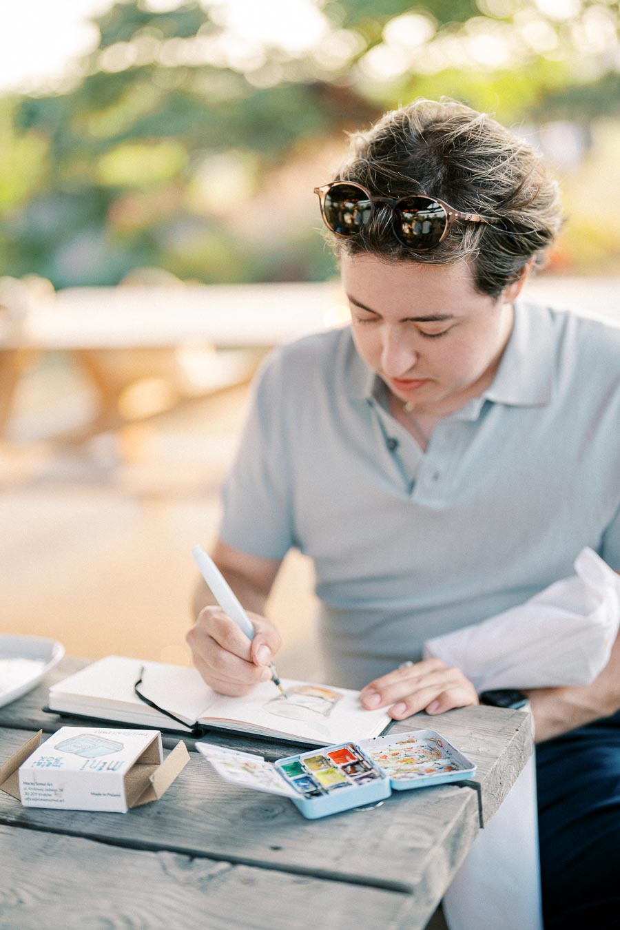 Person sketching with a pen in a notebook at a wooden outdoor table, surrounded by art supplies and paint palette, under soft sunlight.
