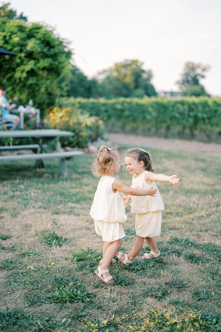 Two young children in matching light-colored outfits joyfully dancing on a grassy field, surrounded by lush greenery and a bench in the background on a sunny day.