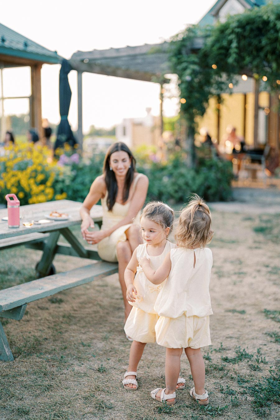 A woman sitting at an outdoor picnic table in a garden setting, smiling at two young children playing nearby, all wearing matching light-colored outfits and surrounded by lush greenery and flowers.