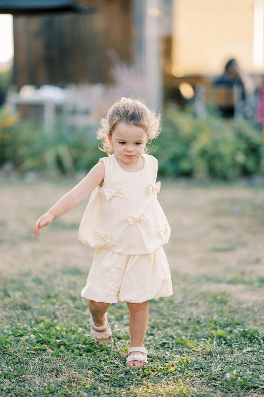 A young girl wearing a light yellow dress with bows, happily running on a grassy field in a sunny outdoor setting.