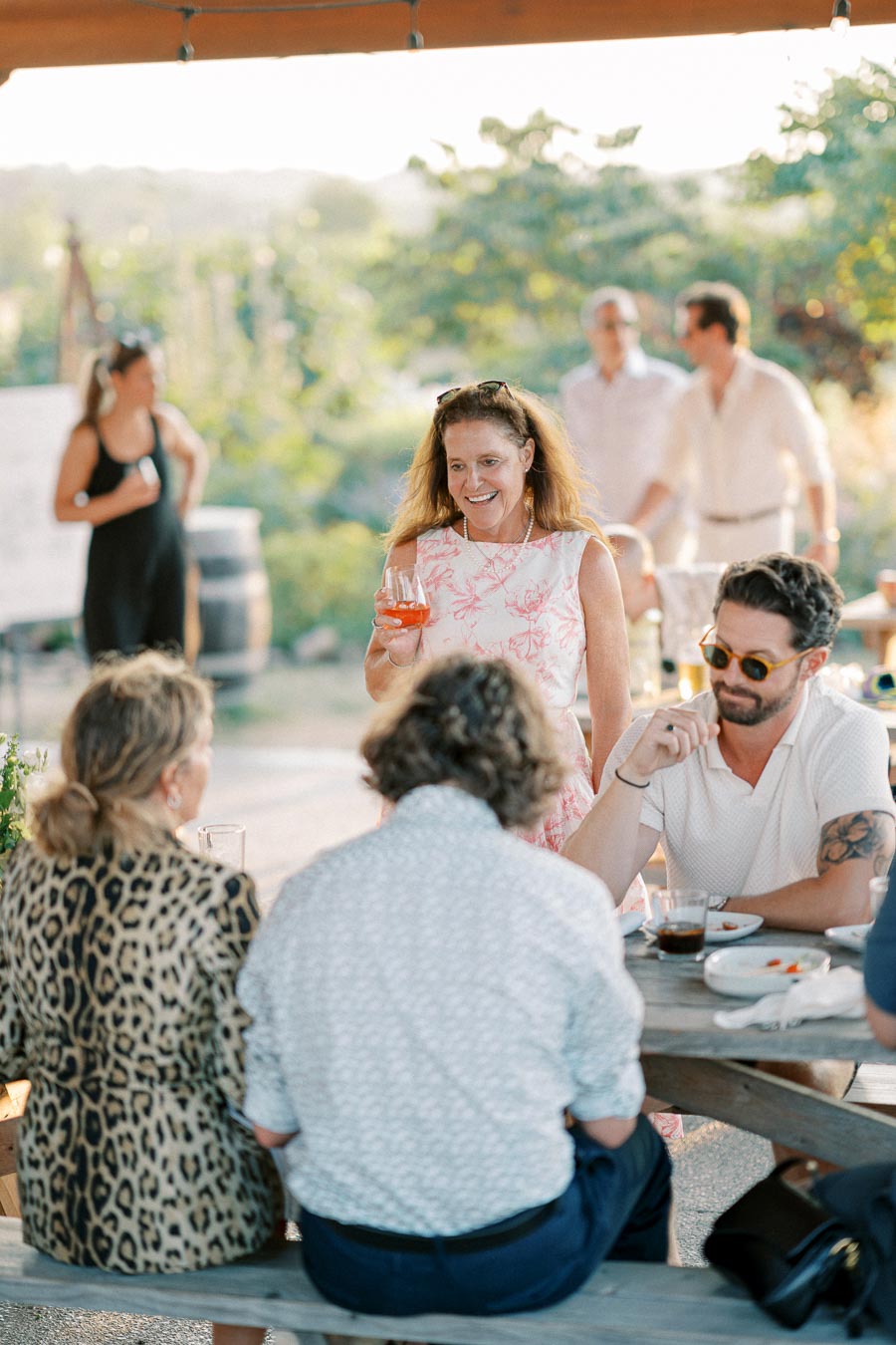A group of people enjoying a casual outdoor gathering, featuring a woman in a floral dress holding a drink and mingling with seated friends at a rustic wooden table in a sunny garden setting.