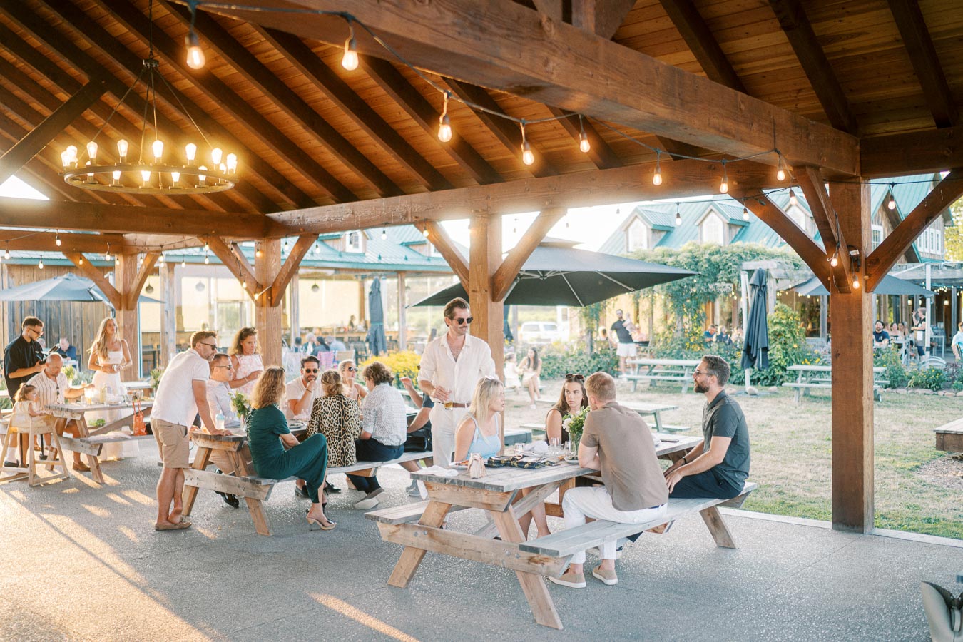 Outdoor gathering under a wooden pavilion with people enjoying food at picnic tables, string and chandelier lights creating a warm, inviting atmosphere.