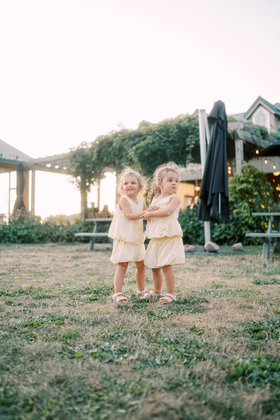 Two young children in matching yellow dresses stand on a grassy area, smiling and holding hands, with a backdrop of a garden and building.