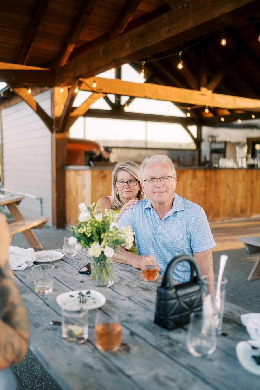 A couple enjoying drinks at an outdoor wooden table, adorned with a vase of fresh flowers, under a rustic pavilion with string lights.