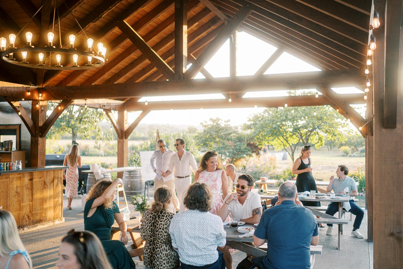 A group of people enjoying a sunny outdoor gathering under a wooden pavilion with string lights, surrounded by trees and fields. Guests are seated at picnic tables, chatting and dining in a relaxed atmosphere.
