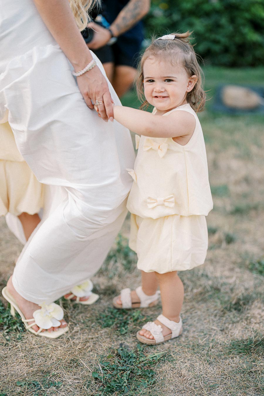 Toddler in a light yellow dress smiling and holding onto an adult's leg outdoors.