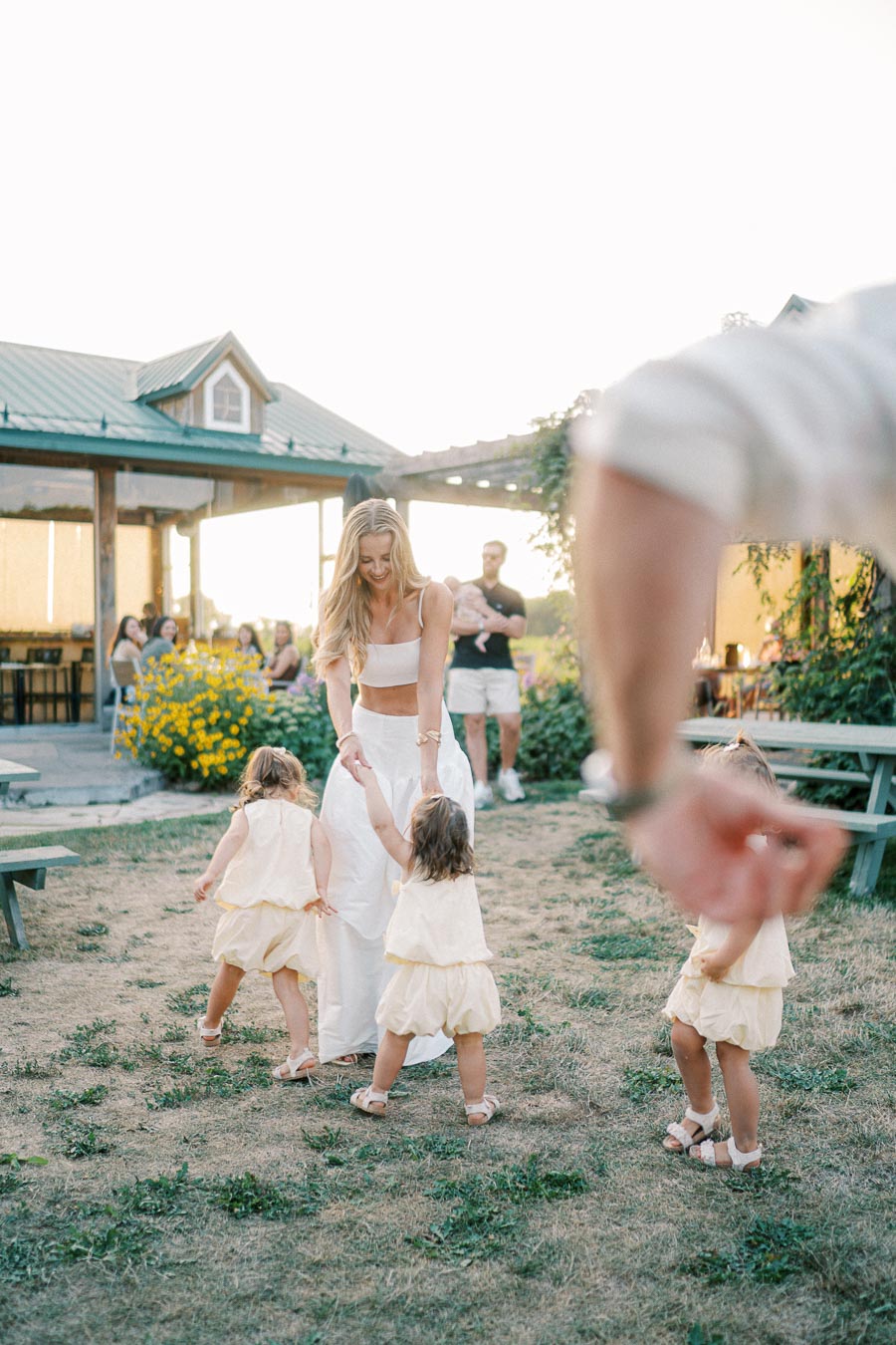 A woman enjoying a sunny outdoor gathering, dancing with two young children in matching yellow outfits, with a rustic pavilion and family in the background.