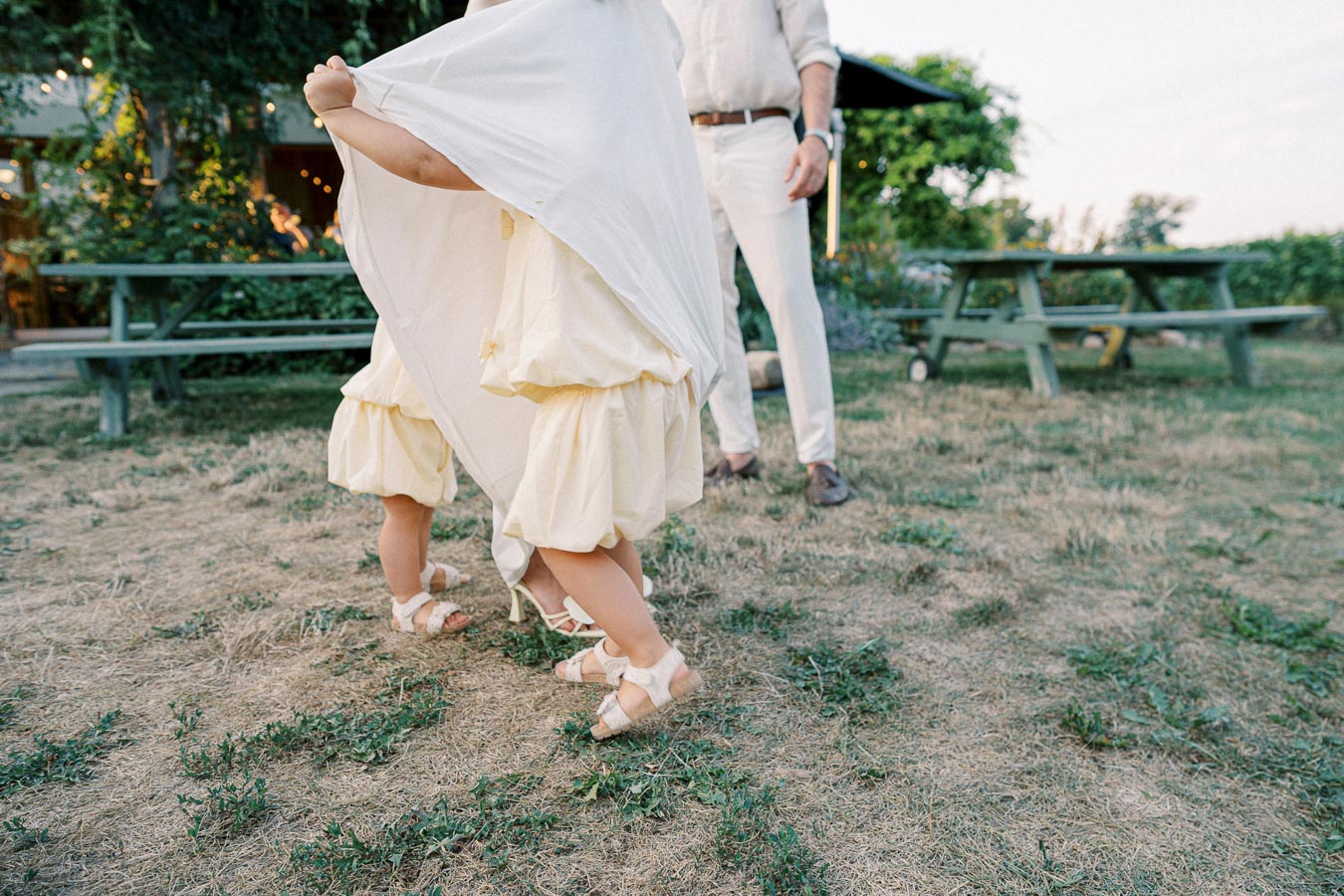 Young children playfully hiding under a white cloth in a garden setting. The scene includes picnic tables and greenery in the background, capturing a joyful outdoor moment.