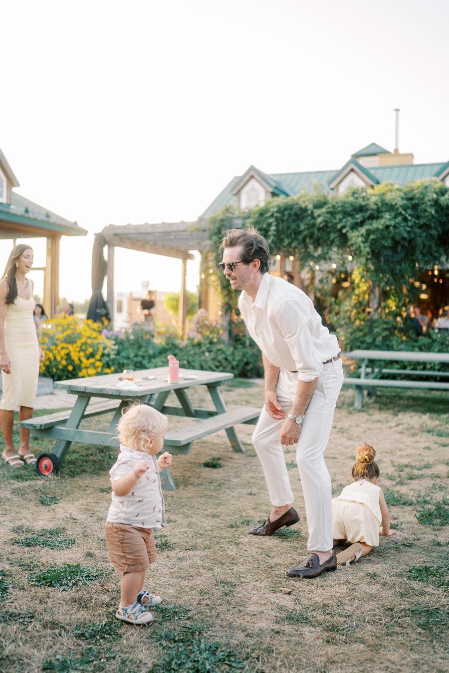 A cheerful outdoor family gathering with adults and children playing on grass near a picnic table, surrounded by greenery and rustic buildings under a clear sky.