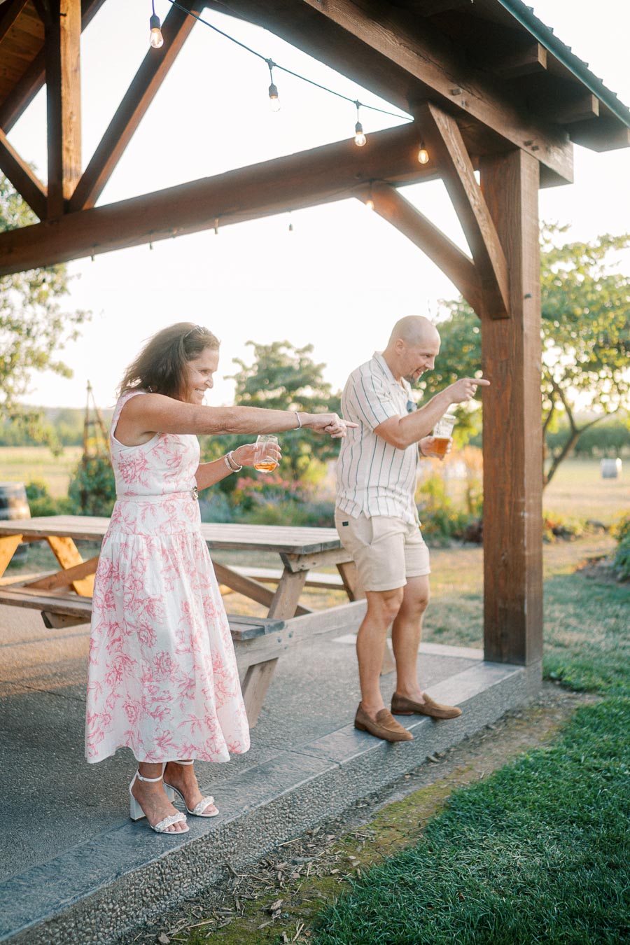 A joyful middle-aged couple dances under a wooden pavilion, enjoying a sunny outdoor setting. The woman wears a floral dress and holds a drink, while the man, in a striped shirt and shorts, mirrors her energy with a smile. The background features lush greenery and warm lighting.