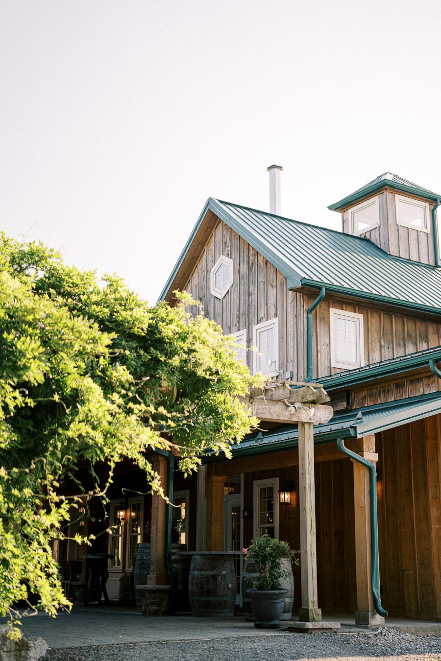 Rustic wooden building with a green metal roof surrounded by lush greenery and wooden barrels, showcasing charming countryside architecture under a clear sky.